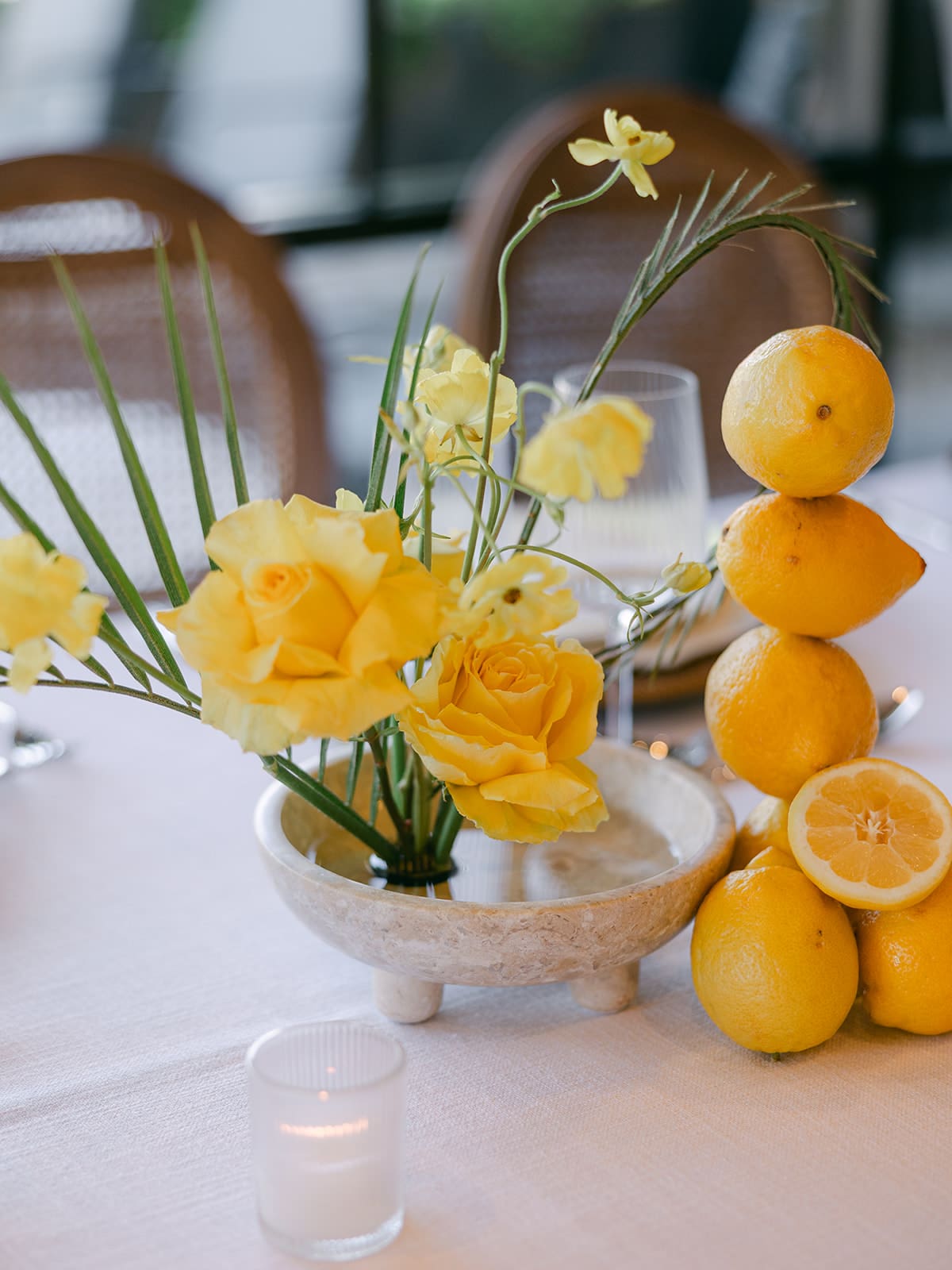 Yellow rose and narcissus floral centerpiece in a travertine footed bowl alongside a stacked lemon tower on a white linen event table