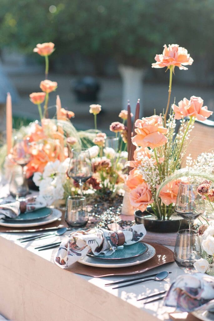 Outdoor wedding tablescape with peach roses, ranunculus, and wispy grasses in low black vessels, styled with smoky glassware and printed napkins