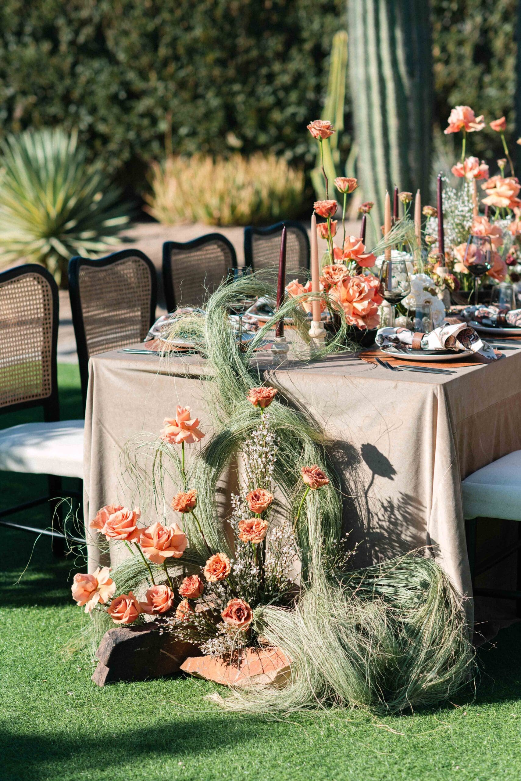 Full outdoor reception table styled with peach roses and trailing grasses, surrounded by saguaro cacti and desert landscaping beneath a clear Arizona sky