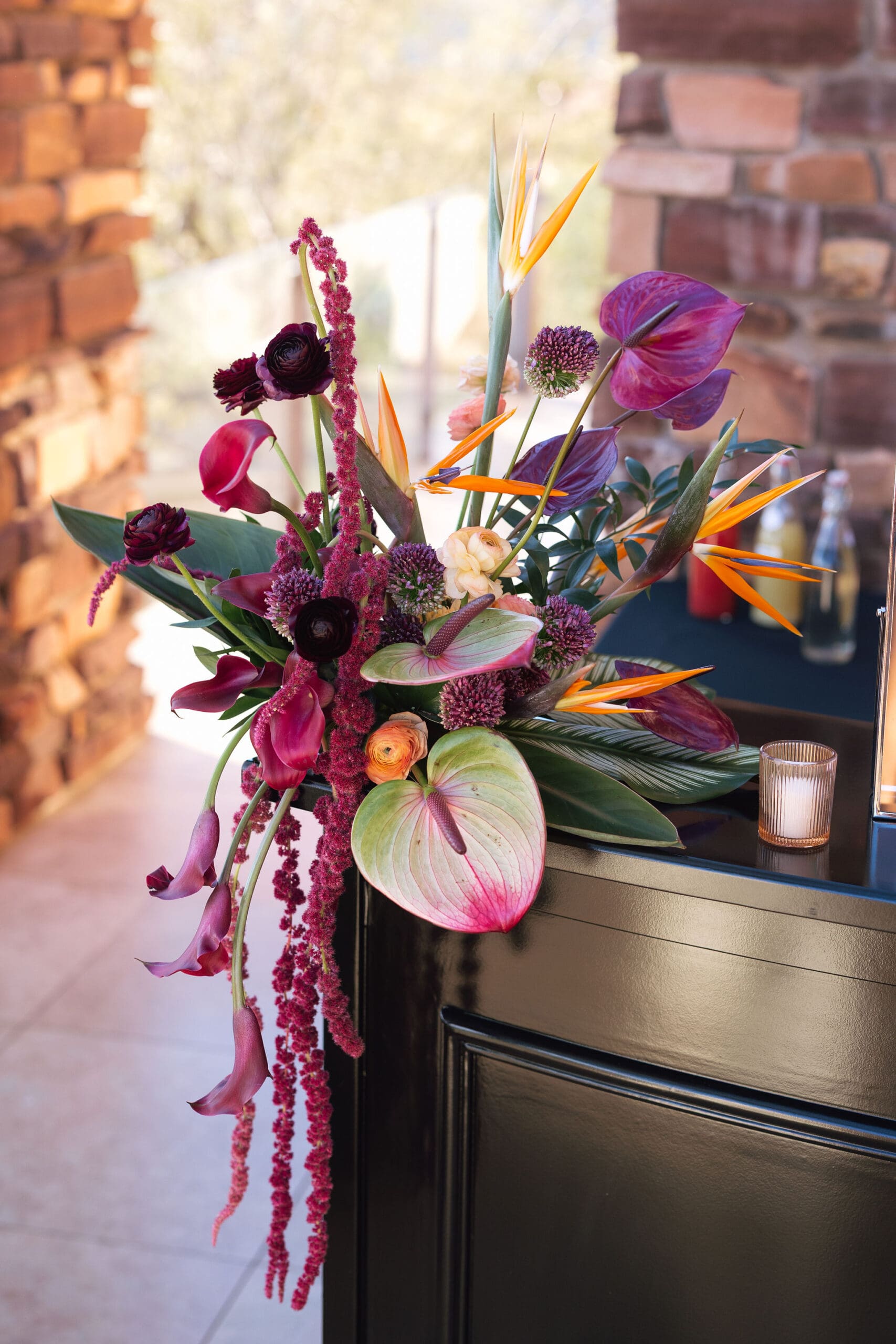 Sculptural floral arrangement featuring bird of paradise, trailing amaranthus, anthurium, dark ranunculus, and calla lilies in deep jewel tones atop a black pedestal