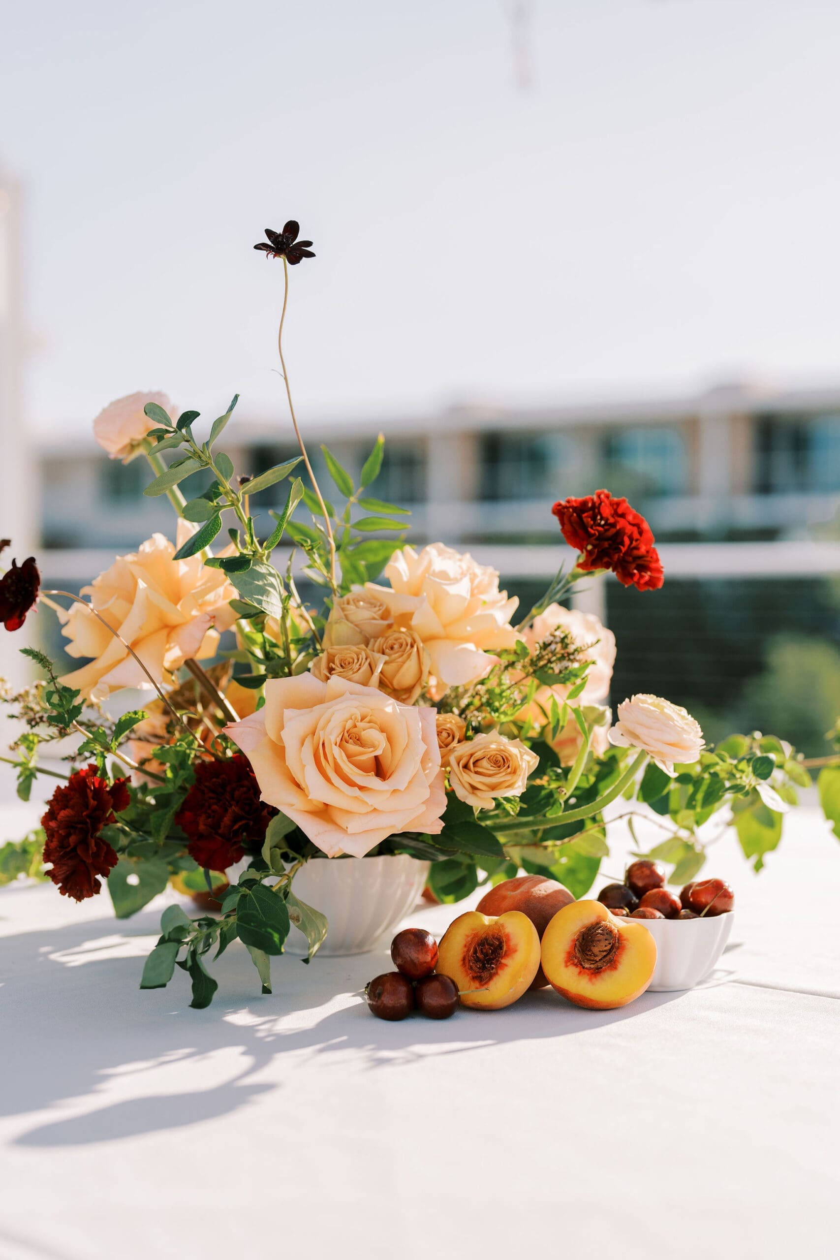 Peach garden rose and dark red ranunculus arrangement in a white bowl with fresh cut peaches and cherries on a white table surface